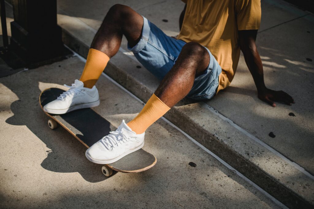 Man wearing denim short and a yellow t-shirt with yellow sock and trainers sitting on the floor with feet resting on a skateboard