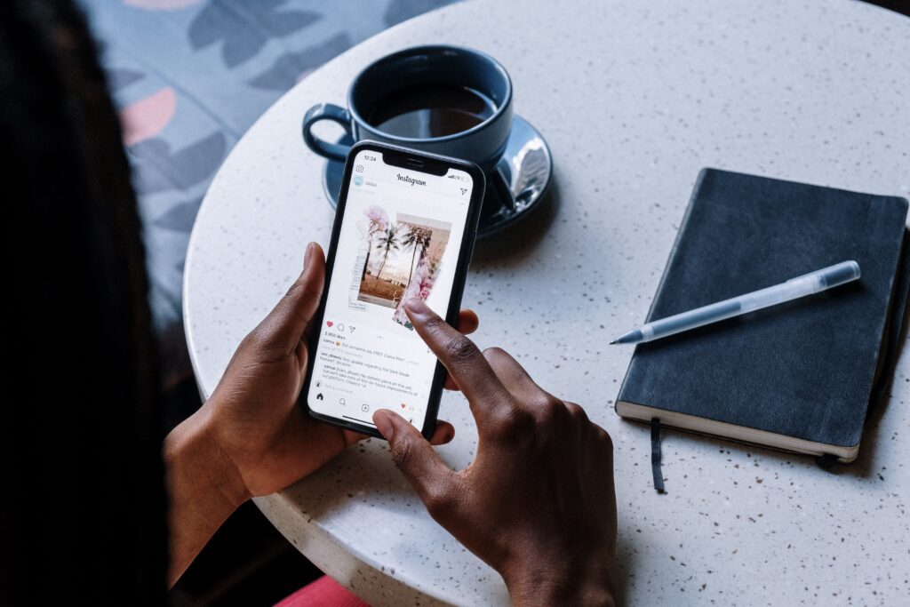 Person Holding Iphone scrolling on social media sat at a table Near Black Ceramic Mug With Coffee and a notepad and pen