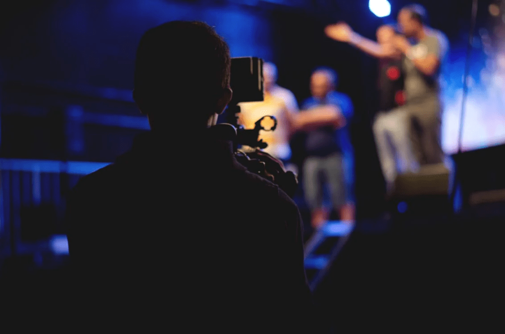 Person holding a camera pointing to people talking on stage.
