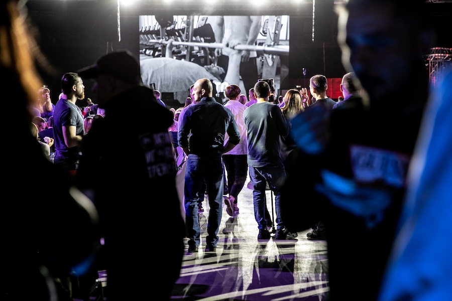 A camera view of an indoor marketing event with dim lighting and a large screen in the displaying a man doing bench presses with someone spotting him. Approximately fifty people are visible in the crowd.