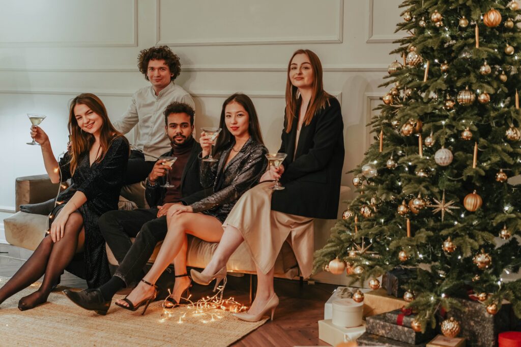 Friends Sitting On A Sofa Beside A Christmas Tree in Cocktail Party Attire holding up a drink