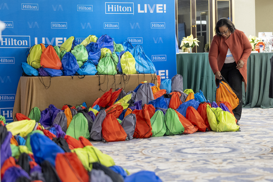 A woman bends over to place a bagged hygiene kit down among a pile of other bagged hygiene kits on a carpeted floor and on a table, with a large "Hilton Live!" wall-to-ceiling poster in the background. This kind of kit-packing activity can be a great way to contribute to ESG goals for meetings.