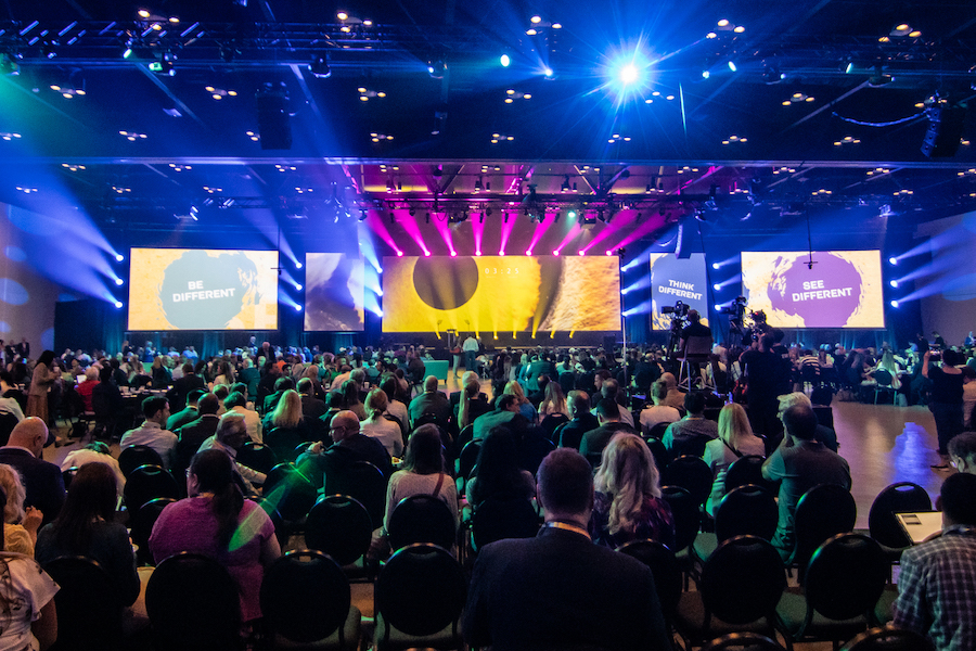 A crowd of several hundred people viewed from behind at the Calgary TELUS Convention Center (CTCC), with five large screens of varying dimensions behind the stage. Lights of pink, blue, purple, and teal shine across the audience.