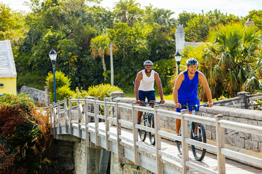 As part of an event theme of sustainability, two men ride on bicycles over a wooden bridge with one of Bermuda's iconic white limestone rooftops visible on a house in the background.