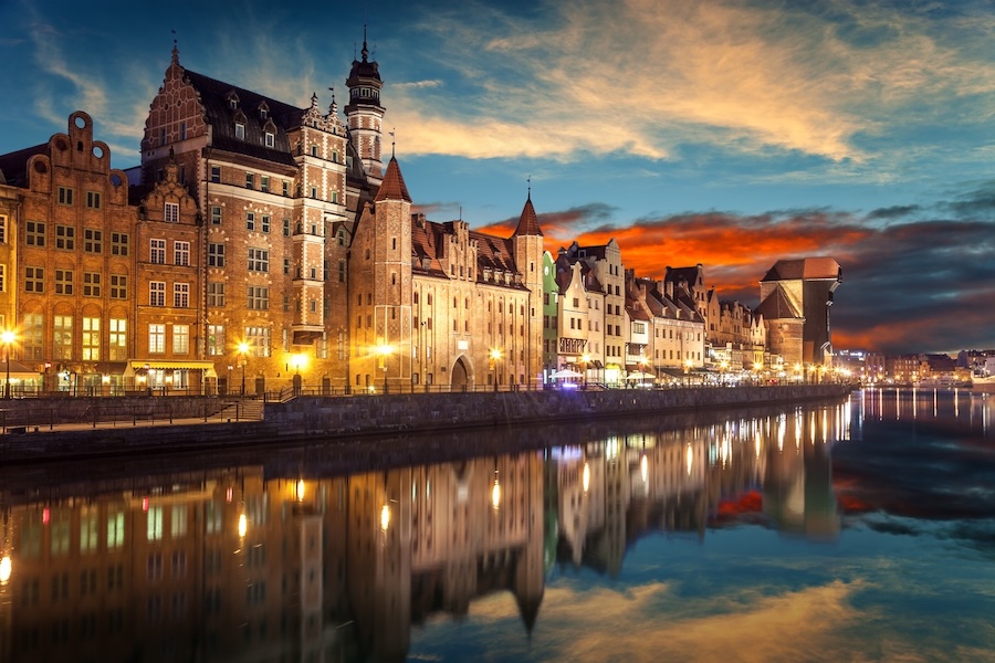 A nighttime riverside view of Gdansk, Poland, with historical architecture and the Crane of Gdansk visible in the background.