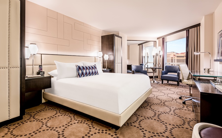 An interior shot of a large king suite at the recently renovated Harrah's Las Vegas. A large bed sits in the middle of a spacious room with sleek, modern decor and a large window facing a panoramic view of the city. Two sofa chairs are visible in the background, along with a work desk on the right-hand side.
