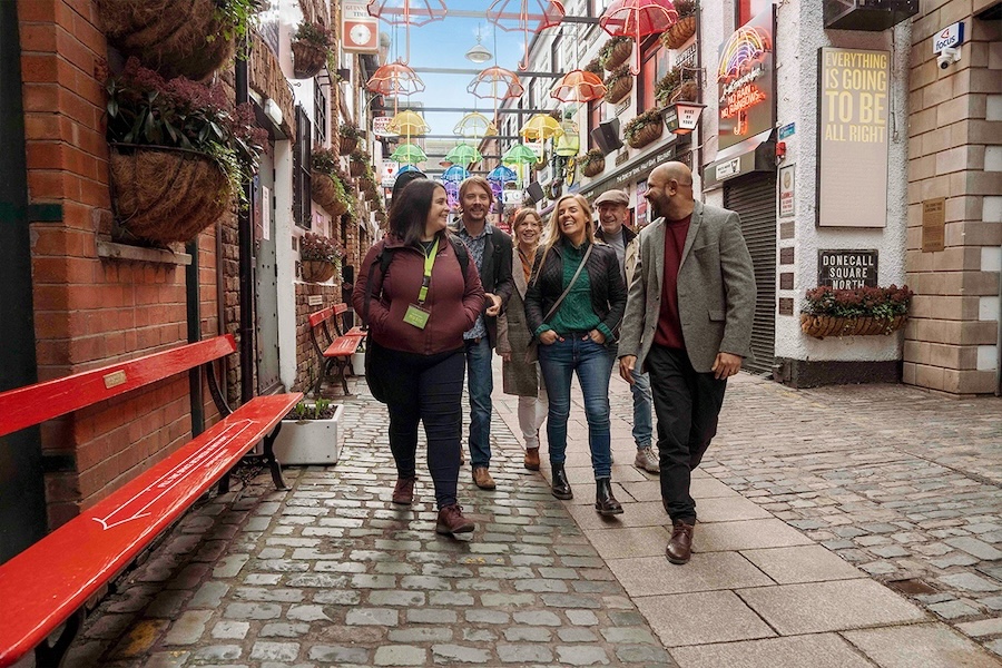Six people chat joyously while walking down the cobblestone street of Music Trail in Belfast. Quaint historical buildings flank both sides, with overhead rainbow colored umbrella sculptures stretching out along the path.