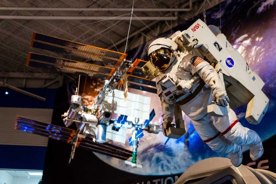 An interior shot of the Space Center Houston, with an authentic astronaut space suit hanging from the ceiling next to satellites, with a large photo of the Earth from space in the background.